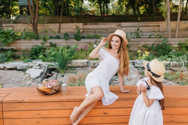 Femme excitée au chapeau vintage posant sur la nature pendant que sa fille la regarde avec intérêt. Portrait en plein air de l'arrière de la petite fille en robe blanche debout à côté du parterre de fleurs avec maman.