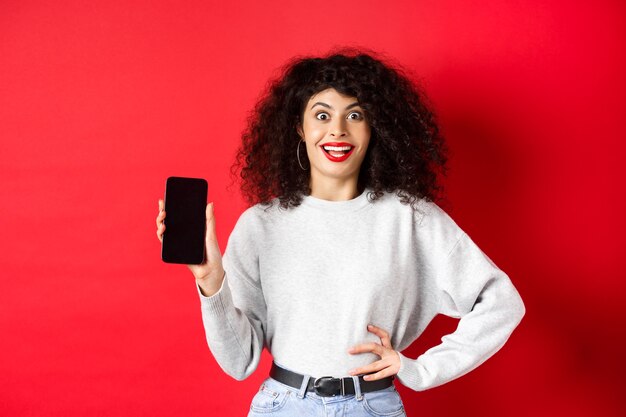 Femme excitée aux cheveux bouclés et aux lèvres rouges, montrant un écran de smartphone vide et criant de joie, debout sur fond rouge.