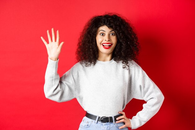 Femme excitée aux cheveux bouclés montrant le numéro cinq avec les doigts, rendant l'ordre, debout sur fond rouge