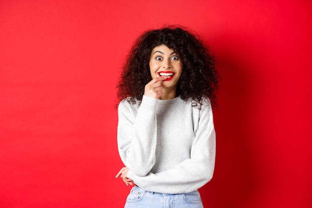 Femme excitée et intriguée désireuse d'essayer quelque chose, faire un regard tenté et se ronger l'ongle amusé, debout sur fond rouge.