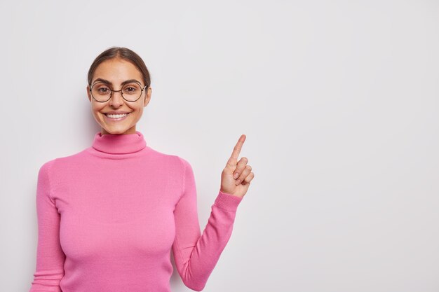 une femme avec une expression joyeuse dit regardez ici indique dans le coin supérieur droit annonce votre texte pose sur blanc porte un col roulé rose et des lunettes rondes