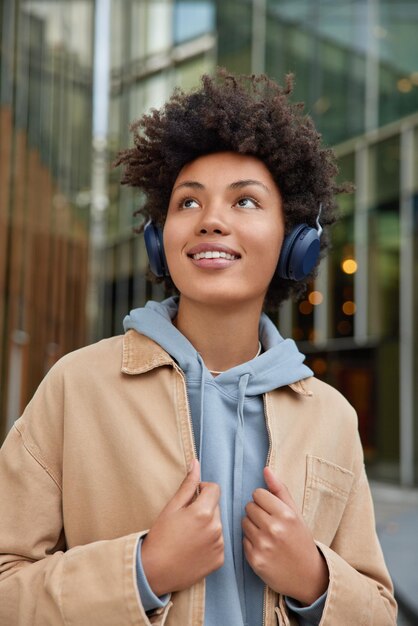 une femme avec une expression joyeuse écoute la piste audio préférée via un casque sans fil porte un sweat à capuche et une veste beige pose à l'extérieur