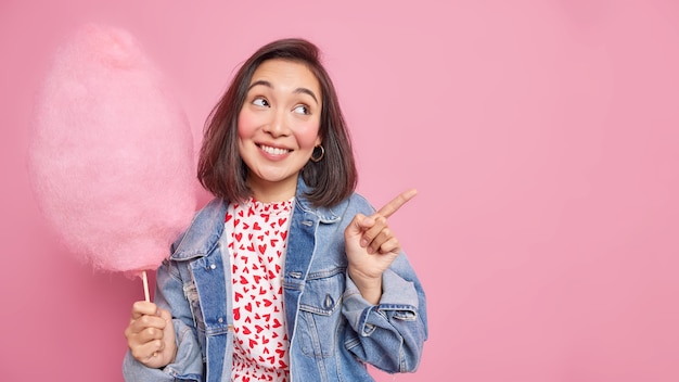 une femme avec une expression joyeuse sourit agréablement indique loin sur l'espace de copie montre la direction détient une délicieuse barbe à papa porte une veste en jean