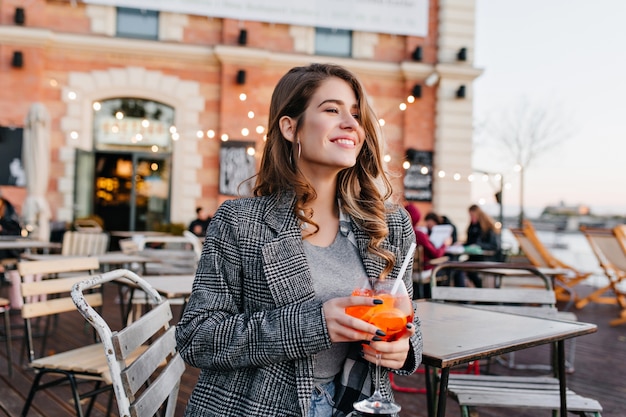 Femme extatique en manteau gris à la voiture avec le sourire tout en buvant une boisson aux fruits au café