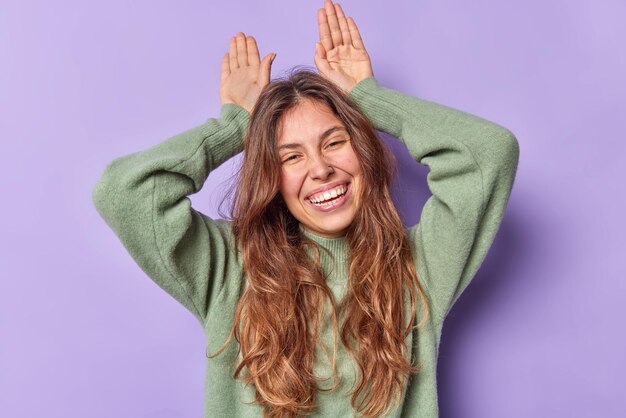 la femme fait des oreilles de lapin avec des paumes sur la tête étant des sourires joyeux et affectueux porte à pleines dents des poses de cavalier décontractées contre le violet amuse l'enfant essaie de vous faire rire