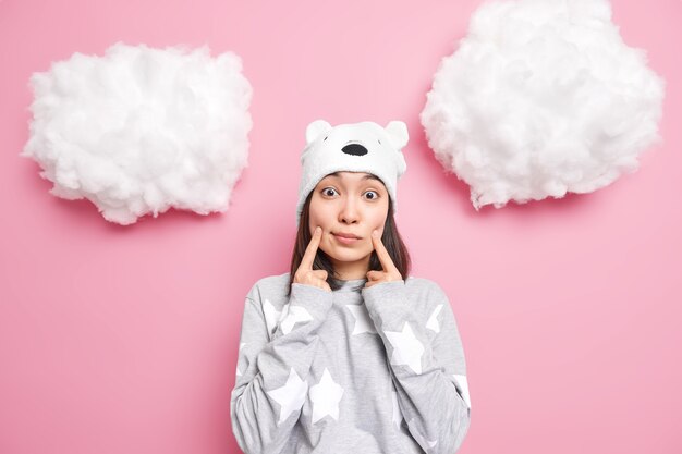 la femme fait des sourires garde les doigts près des coins de la bouche porte un chapeau d'ours doux et un pull décontracté semble isolé sur le rose