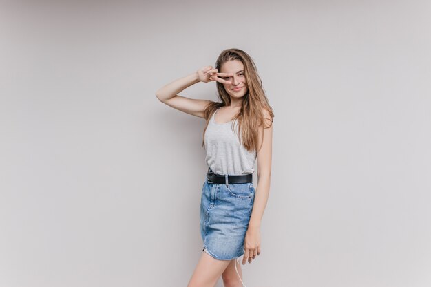 Une femme fascinante porte une jupe en jean et une ceinture noire posant. Portrait intérieur d'une fille adorable aux longs cheveux bruns isolés.