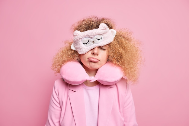 Photo gratuite une femme fatiguée se sent épuisée après un long voyage essaie de faire la sieste en voyageant a une expression endormie porte un masque de sommeil et un oreiller de voyage autour du cou habillé formellement isolé sur un mur rose