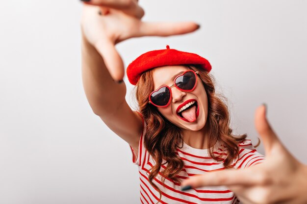 Femme française attrayante à lunettes de soleil en riant à la mode fille au gingembre bouclé s'amuser sur le mur blanc.