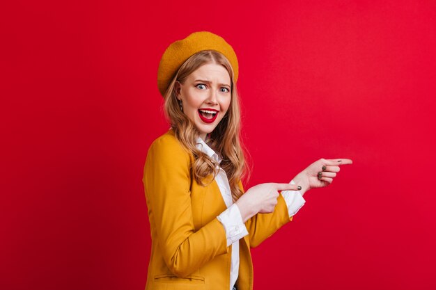 Femme française détendue pointant avec les doigts. fille blonde bien habillée debout sur le mur rouge.