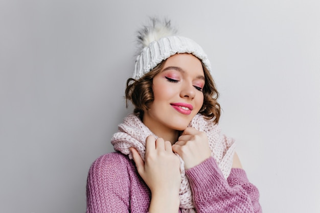 femme frisée blanche heureuse en pull en laine. Portrait intérieur de belle fille avec du maquillage rose posant en chapeau et écharpe en journée froide.