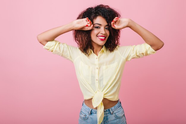 Femme frisée inspirée dans une tenue décontractée à la mode posant avec les mains levées et exprimant vraiment des émotions. Dame africaine sensuelle en jeans dansant de bonne humeur.