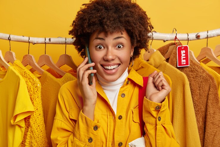 Photo gratuit femme frisée joyeuse avec une expression de visage ravie, un large sourire, lève les poings fermés, téléphone à quelqu'un via un smartphone, porte un sac à provisions sur l'épaule, achète des vêtements.