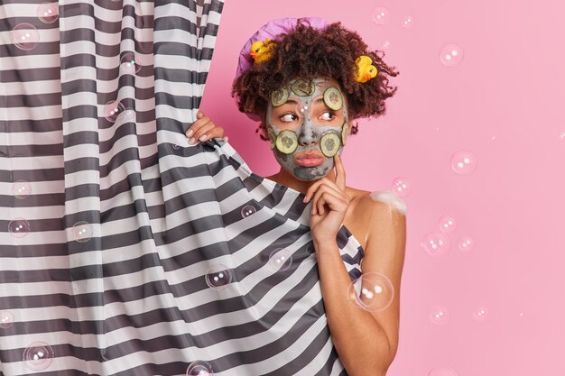 Une femme frisée à la peau sombre réfléchie regarde tristement de côté applique un masque d'argile nourrissant avec des tranches de concombre pour le rajeunissement de la peau se tient nue derrière un rideau de douche isolé sur fond rose