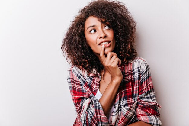 Femme frisée pensif regardant autour et touchant son menton. modèle féminin africain en chemise à carreaux isolé sur la lumière.