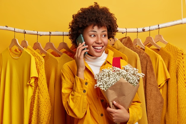 Femme frisée positive a une conversation téléphonique, garde le téléphone portable près de l'oreille, tient un beau bouquet