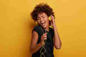 Photo gratuite une femme frisée positive et insouciante entend une superbe piste via un casque, chante à haute voix avec une chanson, tient le smartphone comme microphone