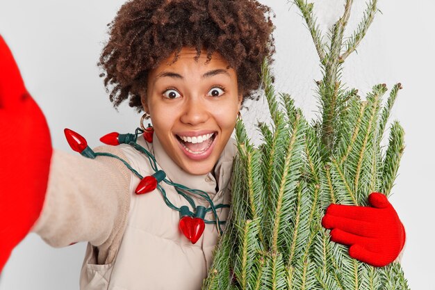 Une femme frisée positive et trop motivée étire le bras, ce qui fait sourire selfie, étant globalement très heureux après avoir acheté un sapin à feuilles persistantes fraîchement coupé pour le Nouvel An se prépare pour des vacances plus savoureuses. Noël approche