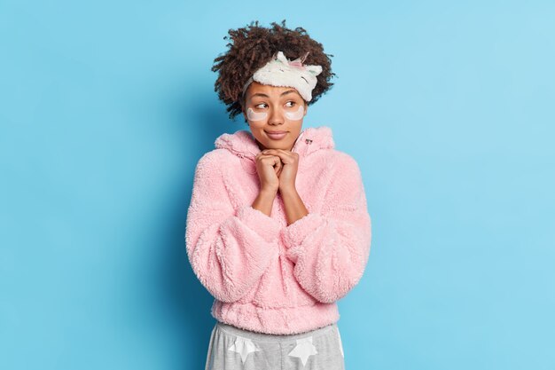 Une femme frisée à la recherche agréable et réfléchie garde les mains sous le menton a des idées imagine quelque chose vêtu de vêtements de nuit applique des coussinets de beauté pour les soins de la peau isolés sur un mur bleu se prépare au sommeil