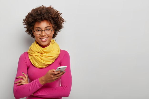 femme frisée à la recherche agréable utilise un téléphone mobile pour discuter en ligne, sourit à pleines dents, porte des lunettes rondes