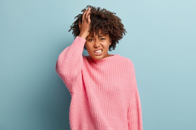 Une femme frustrée malheureuse fronce les sourcils, touche la tête, a les cheveux bouclés, montre des dents blanches, exprime des sentiments négatifs, porte un pull rose tricoté, des modèles à l'intérieur. Lady regrette quelque chose d'insatisfaction