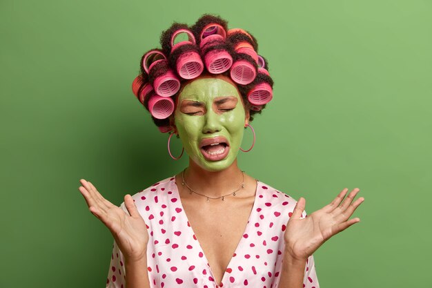 Une femme frustrée et stressante pleure de désespoir, lève la main, découvre une nouvelle tragique, applique un masque facial vert nourrissant, bigoudis, a gâté le week-end, pose à la maison contre le vert