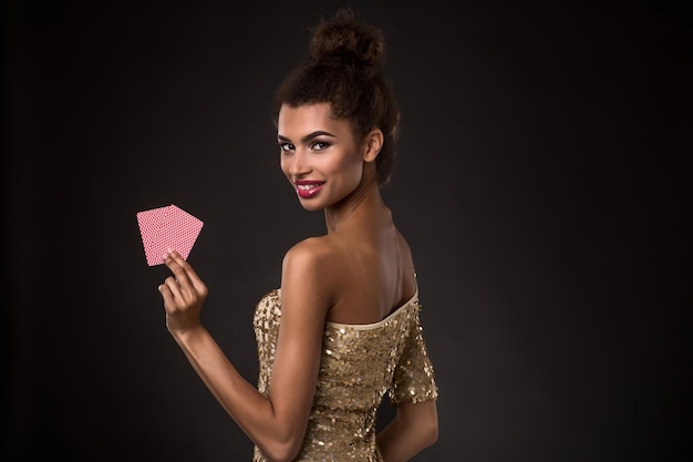 Femme gagnante - Jeune femme vêtue d'une élégante robe dorée tenant deux cartes, une combinaison de cartes de poker d'as. Tourné en studio sur fond noir. Une jeune femme se tient avec son dos