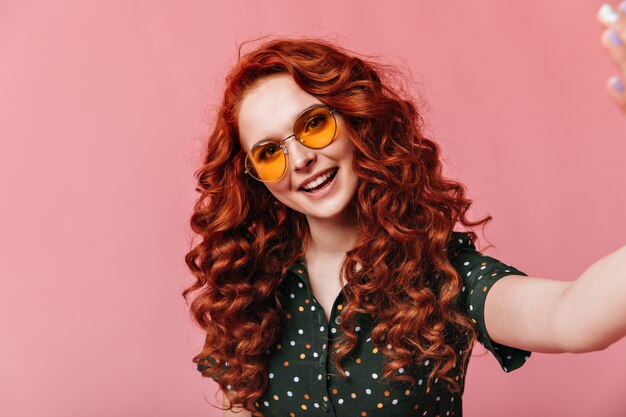 Femme gaie au gingembre prenant selfie. Photo de Studio de curieuse fille bouclée à lunettes de soleil posant sur fond rose.