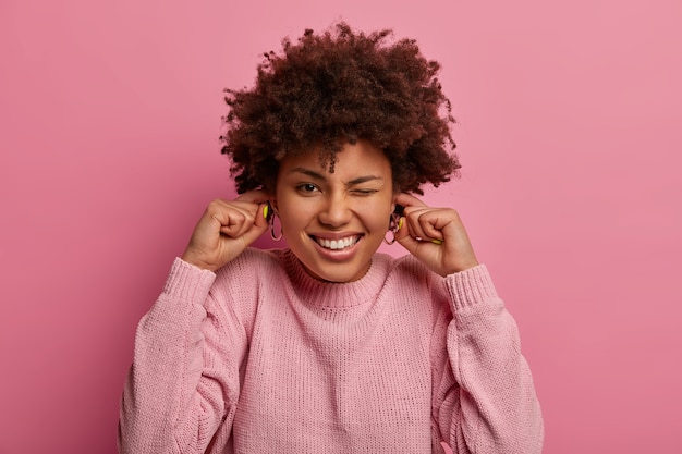 Photo gratuite une femme gaie à la peau sombre et dérangée bouche les oreilles, ignore la musique forte, fait la fête, ne veut pas entendre de bruit, a une coiffure frisée, porte un pull, pose sur un fond rose pastel. décibel