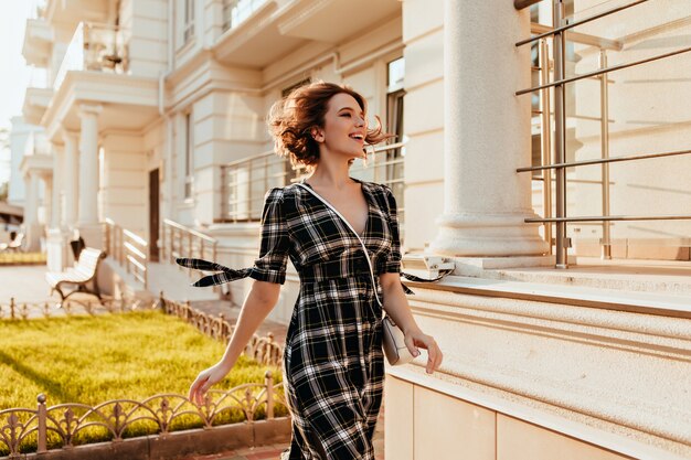 Femme galbée en élégante robe à carreaux marchant dans la rue et riant. Beau modèle féminin blanc avec coupe de cheveux courte, passant du temps à l'extérieur.