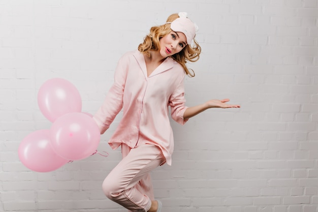 Femme galbée raffinée en pyjama rose dansant avec plaisir sur un mur blanc. Élégante fille d'anniversaire sautant avec des ballons d'hélium et riant.