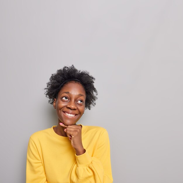 la femme garde la main sous le menton lève les yeux imagine que quelque chose porte un pull jaune décontracté se dresse sur du gris