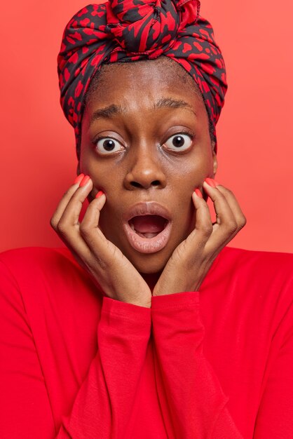 la femme garde les mains sur le visage regarde les yeux obsédés la bouche ouverte regarde avec stupéfaction porte un foulard noué sur la tête pull décontracté isolé sur rouge