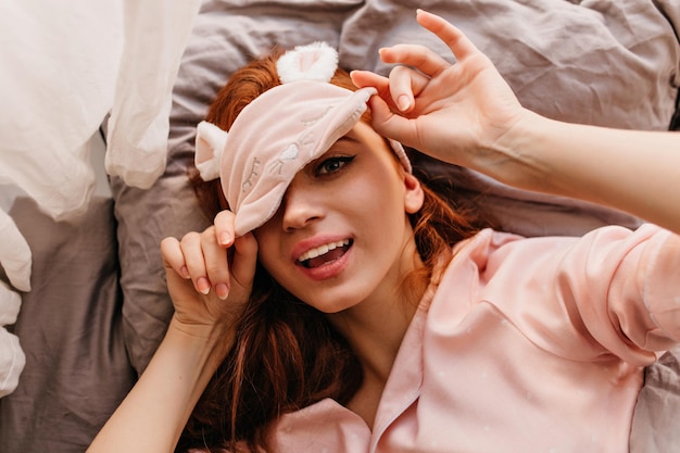 Femme gingembre paresseuse posant dans un masque de sommeil Photo aérienne d'une curieuse fille rousse allongée dans son lit