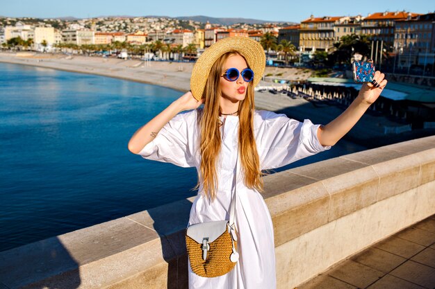 Femme glamour élégante portant une robe blanche de luxe et des accessoires de paille faisant selfie at beach
