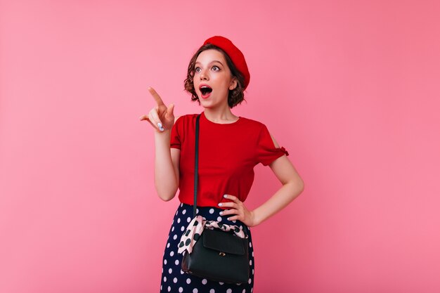 Femme glamour inspirée dans une tenue élégante, pointant le doigt vers quelque chose d'intéressant. Tir intérieur d'une jeune fille caucasienne enthousiaste en béret français et chemisier rouge.