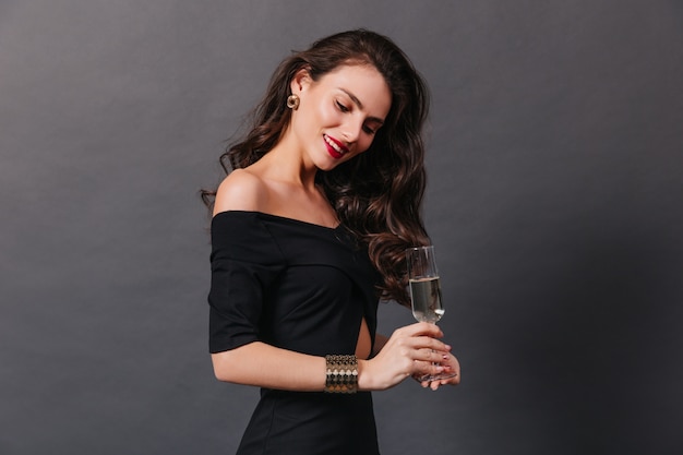 Femme gracieuse aux cheveux longs ondulés et en robe noire élégante posant avec du champagne sur fond sombre.