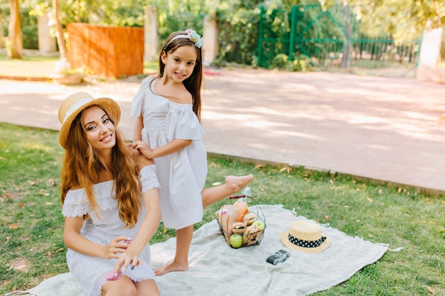 Femme gracieuse en robe blanche assise sur une couverture dans le parc, tandis que sa jolie fille danse derrière son dos. Portrait en plein air de deux soeurs joyeuses s'amusant après le pique-nique.