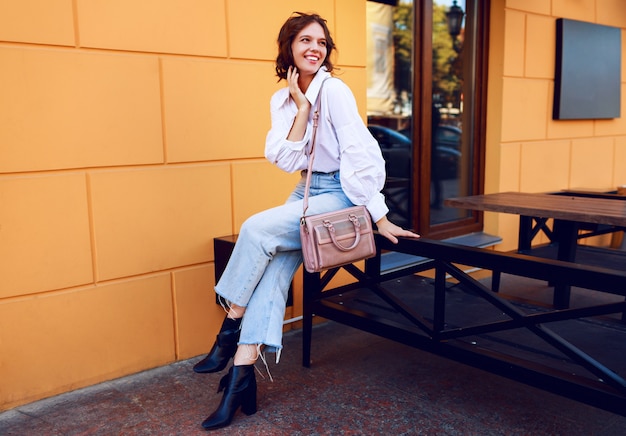 Femme gracieuse en tenue décontractée assise près d'un café moderne aux murs jaunes. Look à la mode. Humeur positive.