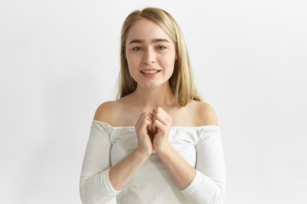 Femme en haut blanc avec les épaules ouvertes tenant les mains sur sa poitrine et souriant avec anticipation