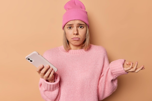 Une femme hésitante, confuse et mécontente, hausse les épaules, semble frustrée devant la caméra, ne sait pas comment effectuer des paiements en ligne, fait défiler les médias sociaux, porte un chapeau rose et un pull isolé sur fond marron