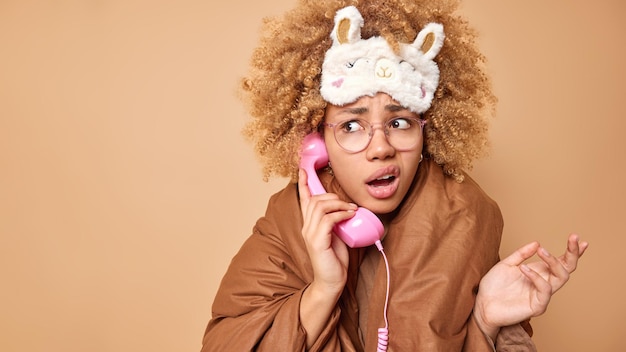 Une femme hésitante se sent mécontente regarde au loin confus hausse les épaules les épaules porte un bandeau sur les yeux enveloppé dans des pourparlers de couverture via le combiné ne se sent pas au courant des poses sur fond marron copiez l'espace pour votre publicité