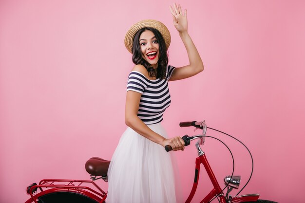 Femme heureuse au chapeau de paille posant à vélo avec un sourire surpris. Plan intérieur d'une jolie fille brune exprimant des émotions heureuses