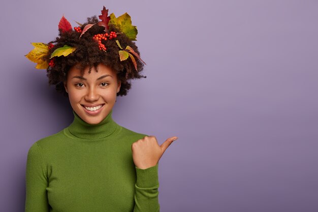 Femme heureuse aux cheveux bouclés décoré par des feuilles d'automne, pose à l'intérieur, pointe le pouce de côté, habillé en col roulé décontracté vert, isolé sur fond violet, sourit largement