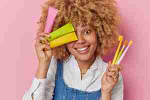 Photo gratuite une femme heureuse aux cheveux bouclés tient des brosses à dents et des tubes de dentifrice sourit avec joie a des dents bien soignées porte une chemise blanche et un jean sarafan isolé sur fond rose concept de soins dentaires