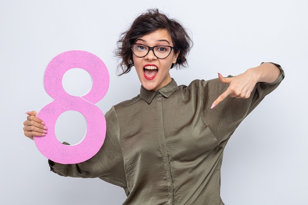 Femme heureuse aux cheveux courts tenant le numéro huit en carton pointant avec l'index vers le bas souriant joyeusement célébrant la journée internationale de la femme le 8 mars debout sur fond blanc