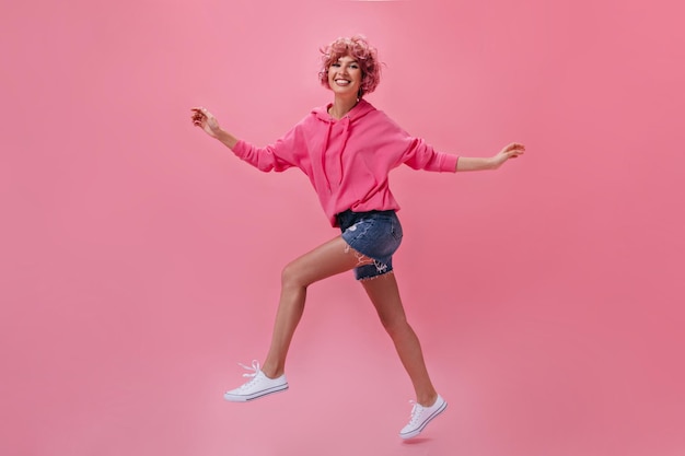 Femme heureuse bouclée en short en jean et sweat à capuche sourit largement et se déplace sur fond rose