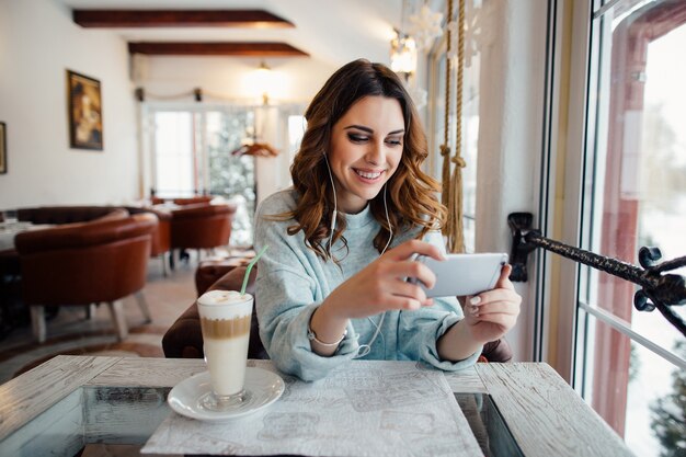 Femme heureuse avec des boucles au café en utilisant un téléphone intelligent et souriant