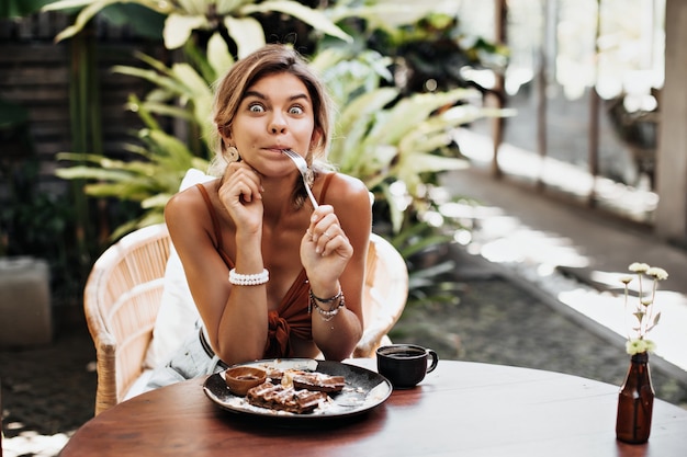 Photo gratuite une femme heureuse bronzée en soutien-gorge brun a l'air surpris, tient une fourchette et fait une grimace