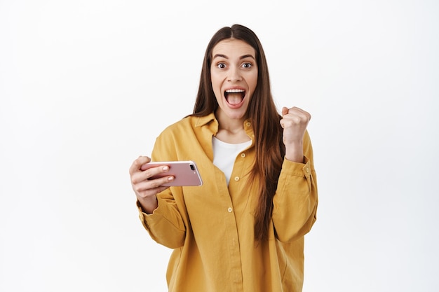 Une femme heureuse crie oui en gagnant en ligne, en atteignant l'objectif, en jouant à un jeu vidéo au téléphone et en célébrant, en faisant pomper le poing et en souriant émerveillée devant, debout sur un mur blanc